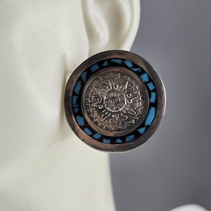May include: A round silver-toned earring featuring a central embossed circular design with intricate patterns. The center is surrounded by a ring of small, inlaid turquoise-colored stone fragments set within a smooth, polished silver-toned metal border.