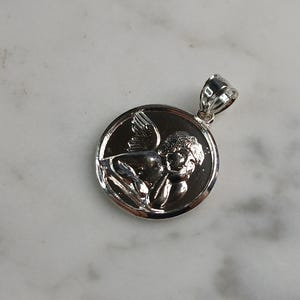 Op de afbeelding: Een zilverkleurige hanger met een gedetailleerd reli&euml;f van een cherubijn met vleugels, geplaatst in een cirkelvormige lijst. De hanger heeft een gepolijste afwerking en een kleine lus voor een ketting. De cherubijn is afgebeeld in een liggende houding.