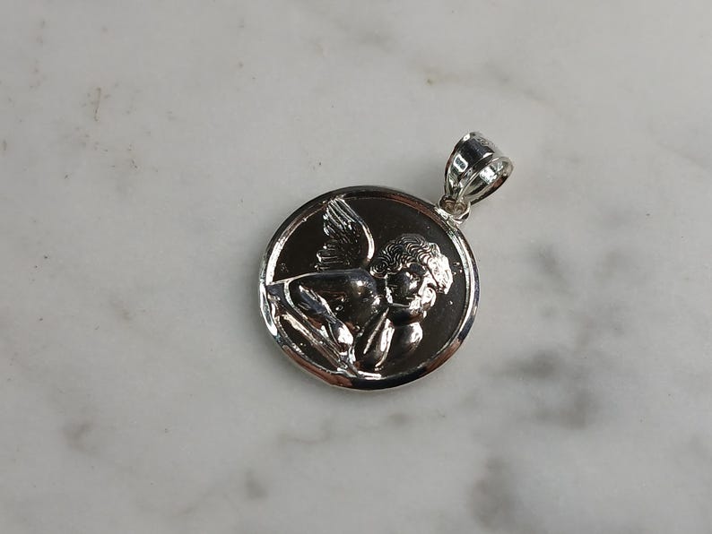 Op de afbeelding: Een zilverkleurige hanger met een gedetailleerd reli&euml;f van een cherubijn met vleugels, geplaatst in een cirkelvormige lijst. De hanger heeft een gepolijste afwerking en een kleine lus voor een ketting. De cherubijn is afgebeeld in een liggende houding.