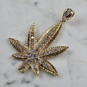 May include: A gold-colored pendant in the shape of a cannabis leaf, encrusted with clear gemstones. The pendant has a textured surface and a loop for a chain. The background is a white and gray marble surface.