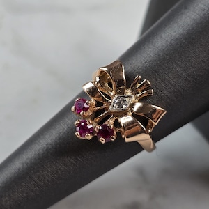 May include: A gold ring featuring a decorative bow design. The bow is accented with three small, round, deep red rubies clustered on one side and a single, small, clear, round-cut diamond set within a marquise-shaped frame at the center.