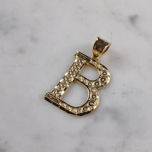 May include: A gold-colored pendant in the shape of the letter "B". The pendant has a textured surface with a pattern of small circles and lines. The bail is a simple loop for attaching to a chain. The pendant is displayed on a white marble surface.