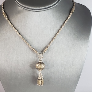May include: A silver-toned necklace with a twisted rope chain and a decorative pendant. The pendant features a floral design on a round orb, with dangling tassels. The necklace is displayed on a gray surface.