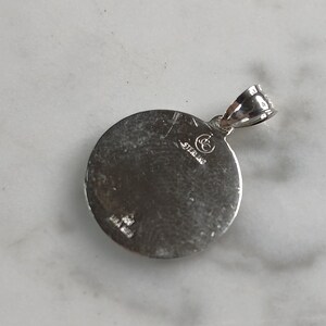 Op de afbeelding: Een ronde, zilverkleurige hanger met een lus voor een ketting. De hanger heeft een getextureerd oppervlak en het woord "STERLING" is erop gestempeld. De hanger ligt op een wit oppervlak.