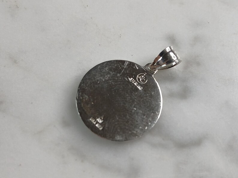 Op de afbeelding: Een ronde, zilverkleurige hanger met een lus voor een ketting. De hanger heeft een getextureerd oppervlak en het woord "STERLING" is erop gestempeld. De hanger ligt op een wit oppervlak.