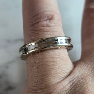 May include: A two-tone ring with a gold band and a silver-coloured centre section. The ring is on a finger, and the design includes small, evenly spaced details around the band. The ring's surface reflects light, highlighting its polished finish.