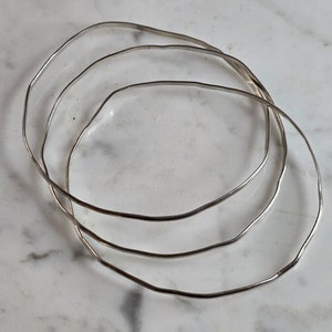 May include: Three silver-colored, wavy-edged bangles are arranged on a marble surface. The bangles are thin and have a slightly irregular shape, creating a unique, handcrafted appearance. The jewelry is simple and elegant.