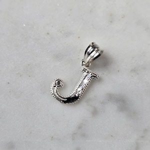 May include: A silver-colored pendant in the shape of the letter "J". The pendant has a decorative design with a loop at the top for a chain. The letter is detailed with a textured border and a dark inlay.
