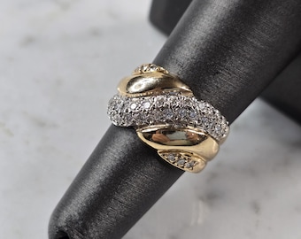 Puede incluir: Un anillo de oro y diamantes con un diseño único. El anillo presenta un patrón arremolinado con una banda de pequeños diamantes engastados entre secciones de oro. El anillo se muestra sobre una superficie negra.