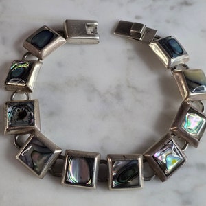 May include: A silver bracelet with a clasp closure features a series of square-shaped abalone shell inlays. The shell inlays have a variety of iridescent colors, including blue, green, and purple.