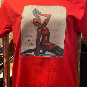 May include: Red t-shirt with a graphic print of a person holding a seashell. The text "Nou se Libète" is printed below the image.