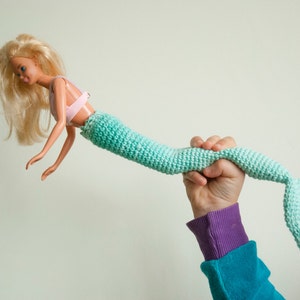 May include: A hand holds a crocheted mermaid tail, attached to a Barbie doll. The tail is a light blue and green ombre color.