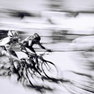 May include: Black and white image of two cyclists in a race, captured with a motion blur effect. The cyclists wear helmets and cycling gear, riding sleek road bikes. The background is blurred, emphasizing speed.