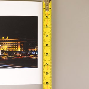 May include: A photograph of a nighttime cityscape with a yellow measuring tape. The cityscape features a building with a green roof and yellow lights. The measuring tape shows a measurement of approximately 8 inches.