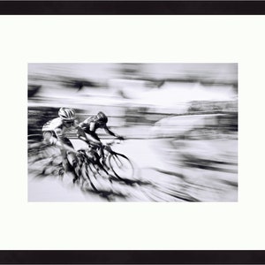 May include: Black and white framed photograph of two cyclists in a race, captured with a motion blur effect. The cyclists are wearing helmets and riding bicycles. The image is set against a white mat and framed in a black frame.