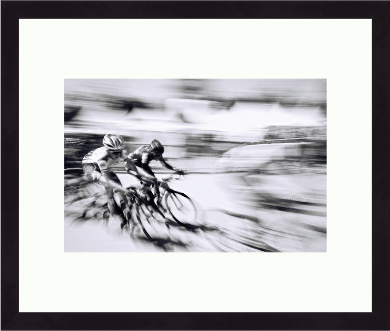 May include: Black and white framed photograph of two cyclists in a race, captured with a motion blur effect. The cyclists are wearing helmets and riding bicycles. The image is set against a white mat and framed in a black frame.