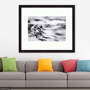 May include: Black and white framed print of cyclists in motion. The image captures the blurred movement of two cyclists racing, creating a sense of speed and dynamism. The artwork is displayed above a gray sofa with colorful throw pillows.