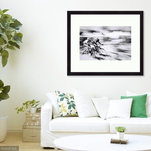 May include: Black and white framed print of cyclists in motion. The artwork captures the speed and energy of a cycling race, with blurred lines suggesting movement. The frame is black, and the print is displayed in a living room setting.