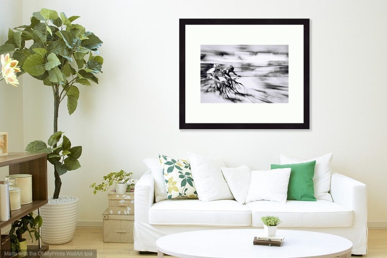 May include: Black and white framed print of cyclists in motion. The artwork captures the speed and energy of a cycling race, with blurred lines suggesting movement. The frame is black, and the print is displayed in a living room setting.