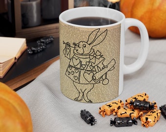 May include: White ceramic mug with a black and white illustration of the White Rabbit from Alice in Wonderland holding a pocket watch and a scroll. The mug is filled with coffee and has a black handle. The mug is sitting on a table with a white tablecloth and some orange and black candy.