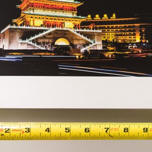 May include: A photograph of a Chinese pagoda at night, illuminated with warm yellow and orange lights. The image captures the building's intricate architecture and the surrounding cityscape, with streaks of light from passing vehicles.