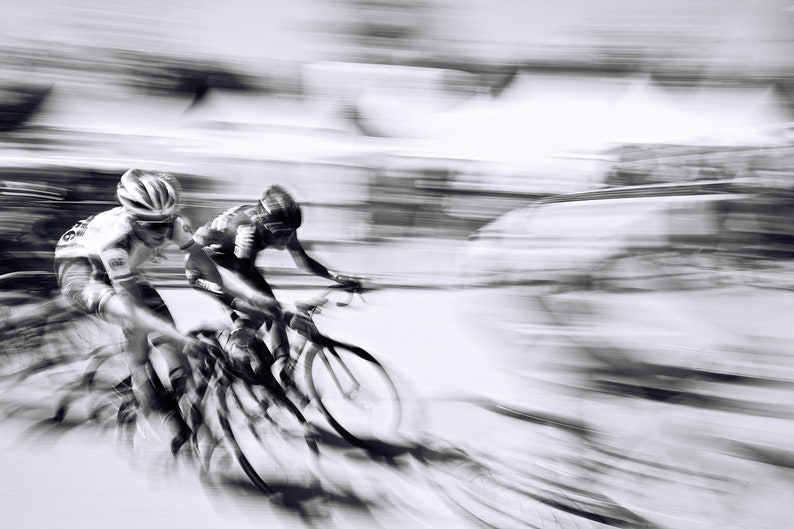 May include: Black and white image of two cyclists in a race, captured with a motion blur effect. The cyclists wear helmets and cycling gear, riding sleek road bikes. The background is blurred, emphasizing speed.