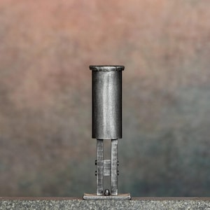 Könnte beinhalten: Ein handgeschmiedetes Metallwerkzeug mit einem zylindrischen Oberteil, das an einem vertikalen Rahmen mit zwei parallelen Stangen und einem kleinen kugelförmigen Akzent an der Basis befestigt ist. Das Objekt hat eine dunkle, texturierte Oberfläche.