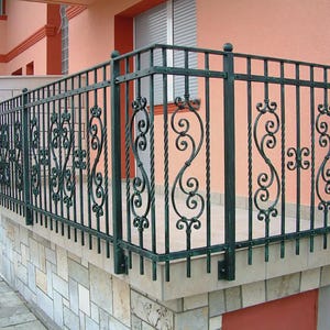 Puede incluir: Una barandilla de balcón de hierro forjado verde oscuro con adornos decorativos y barras verticales. La barandilla está montada sobre una base de piedra y se encuentra contra un exterior de edificio color melocotón.