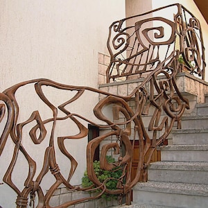 May include: Ornate, brown metal railing with an abstract design. The railing features swirling, organic shapes and is installed on a set of concrete stairs. The design is repeated on a smaller section of railing above the stairs.