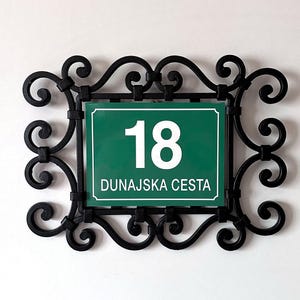 以下が含まれることがあります： 白い数字の18と白い文字でDUNAJSKA CESTAと書かれた長方形の緑色の番地プレート。プレートは、装飾的なスクロール模様と曲線を描く鉄のアクセントが施された黒い金属製のフレームに収められています。