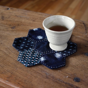 May include: A white ceramic teacup filled with tea sits on a navy blue and white patterned hexagonal coaster. The coaster is made of fabric and has white stitching. The cup and coaster are on a wooden surface.