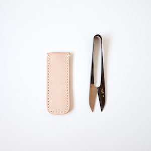 May include: A pair of black and silver scissors with a light tan leather sheath. The scissors have a curved handle and sharp blades. The sheath is rectangular and stitched along the edges.