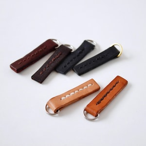 May include: Five leather keychains with different stitching patterns and colors. The keychains are brown, black, and tan. Each keychain has a metal ring.