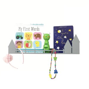 May include: A gray metal shelf shaped like a fence and houses, holding a children's book titled "My First Words", a green toy, and a book with Italian text. A beaded necklace and a pink headband hang from the shelf.