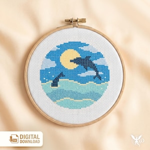 May include: A cross-stitch embroidery hoop depicting a dolphin leaping from the sea under a full moon. The design uses blue, white, and yellow. The text "DIGITAL DOWNLOAD" is in the lower left corner. The hoop is held in a wooden frame.