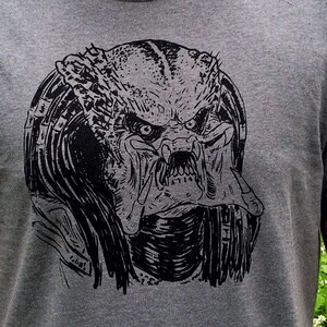 May include: A black and white graphic print of a Predator's head on a gray t-shirt.