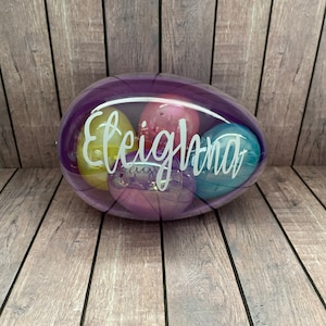May include: A clear plastic egg-shaped container with a purple tint, filled with colorful plastic eggs. The container has the name "Eleighna" written on it in white.