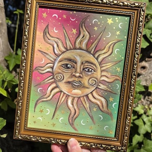 May include: A framed painting featuring a central sun face with golden rays, set against a gradient background transitioning from pink to green. The artwork is decorated with small white crescent moons and yellow stars, enclosed in an ornate gold frame.