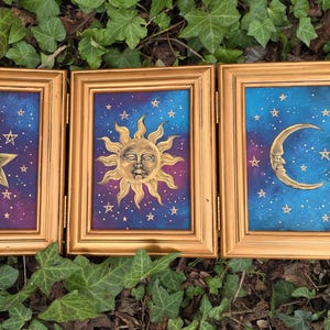 May include: A triptych of three gold-toned frames containing paintings of celestial bodies. The left frame features a star with a face, the centre a sun with a face, and the right a crescent moon with a face, all set against a blue and purple starry sky.