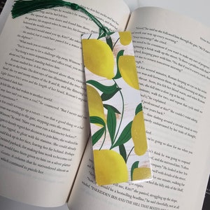 May include: A close-up of a bookmark with a lemon and leaf design. The bookmark is white with a yellow lemon and green leaves. The text "INKRIDDEN IRIS AND THE HILL THAT BESTED HER." is printed on the bookmark.