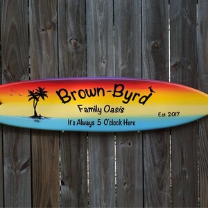 Könnte beinhalten: Ein surfbrettförmiges Holzschild mit einem Regenbogenverlauf. Das Schild trägt den Text "Brown-Byrd Family Oasis", "It's Always 5 O'clock Here" und "Est. 2017". Eine Silhouette einer Palme ist ebenfalls abgebildet.