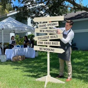 May include: A wooden signpost with directional arrows, displaying event details: "GRAND OPENING!", "NEW PRODUCTS", "EXCLUSIVE DEALS", "FRIE SWAG BAGS", and "NETWORKING". A person is positioned near the sign.