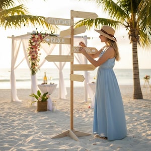 May include: A wooden directional signpost with arrows pointing to "Ceremony", "Beach", and "Wedding" on a sandy beach. A woman in a light blue dress and a straw hat is adjusting the sign. The scene is set for a beach wedding.