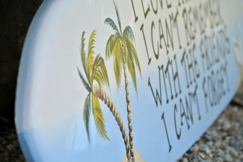 May include: Oval wooden sign with a light blue and white background, featuring two palm trees and the text "I LOVE THE MEMORIES I CAN'T REMEMBER WITH THE FRIENDS I CAN'T FORGET".