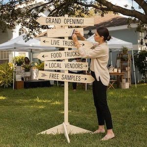 Könnte beinhalten: Ein hölzernes Wegweiser mit Pfeilen, die auf "GRAND OPENING!", "LIVE MUSIC", "FOOD TRUCKS", "LOCAL VENDORS" und "FREE SAMPLES" zeigen. Das Schild steht auf einer Wiese, eine Person justiert es. Zelte und ein Haus sind im Hintergrund zu sehen.