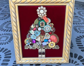 May include: A framed Christmas tree artwork made from vintage jewelry. The tree is composed of various brooches, earrings, and other embellishments in shades of gold, silver, red, blue, and green. The background is a deep red velvet, and the frame is gold.