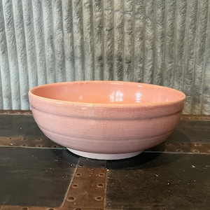 May include: A pale pink ceramic bowl with a slightly textured surface. The bowl is round and has a wide, shallow shape.