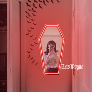 May include: A red neon coffin-shaped mirror with a woman reflected in it. The mirror is on a pink wall with black bats decorating the wall. The text "Arts Vogue" is written in black on the wall.