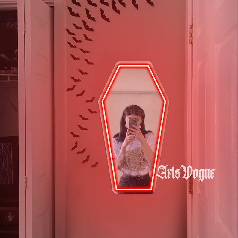 May include: A red neon coffin-shaped mirror with a woman reflected in it. The mirror is on a pink wall with black bats decorating the wall. The text "Arts Vogue" is written in black on the wall.
