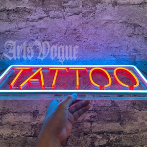 May include: A neon sign with the word "TATTOO" in red neon letters on a clear glass background. The sign is mounted on a brick wall.